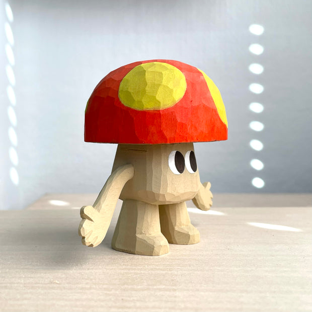 Whittled wooden sculpture of a mushroom with arms and large cartoon eyes and no other facial features. It's body is the exposed natural wood color and its cap is red with large yellow spots.