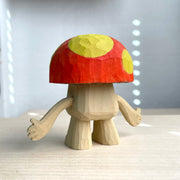 Whittled wooden sculpture of a mushroom with arms and large cartoon eyes and no other facial features. It's body is the exposed natural wood color and its cap is red with large yellow spots.