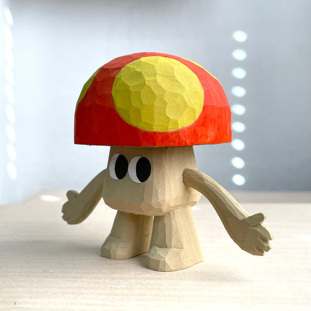 Whittled wooden sculpture of a mushroom with arms and large cartoon eyes and no other facial features. It's body is the exposed natural wood color and its cap is red with large yellow spots.