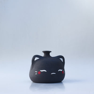 Small, short black jar with a painted on cat face, with closed eyes and red cheeks. It has a small spout opening.