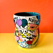 Ceramic vessel with many colorful doodles all around it. Thematics are: smiling fruit, rainbows, clouds, balloons, and various exclamations. Interior of the vessel is a teal blue.