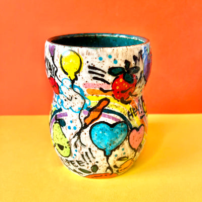 Ceramic vessel with many colorful doodles all around it. Thematics are: smiling fruit, rainbows, clouds, balloons, and various exclamations. Interior of the vessel is a teal blue.