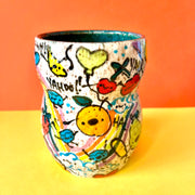 Ceramic vessel with many colorful doodles all around it. Thematics are: smiling fruit, rainbows, clouds, balloons, and various exclamations. Interior of the vessel is a teal blue.