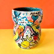 Ceramic vessel with many colorful doodles all around it. Thematics are: smiling fruit, rainbows, clouds, balloons, and various exclamations. Interior of the vessel is a teal blue.