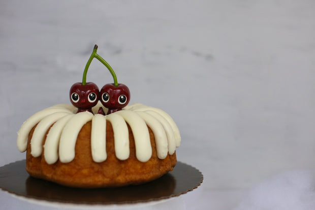 Sculpture of a pair of cherry characters, with large cute eyes and small mouths. They press their palms against one another and their heads are attached to the same stem. They sit in a bundt cake.