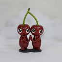 Sculpture of a pair of cherry characters, with large cute eyes and small mouths. They press their palms against one another and their heads are attached to the same stem.