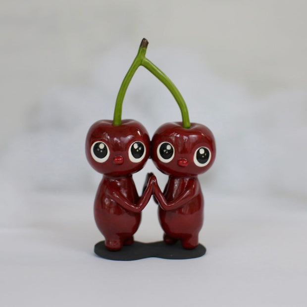 Sculpture of a pair of cherry characters, with large cute eyes and small mouths. They press their palms against one another and their heads are attached to the same stem.