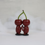 Sculpture of a pair of cherry characters, with large cute eyes and small mouths. They press their palms against one another and their heads are attached to the same stem.