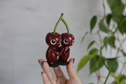 Sculpture of a pair of cherry characters, with large cute eyes and small mouths. They press their palms against one another and their heads are attached to the same stem.