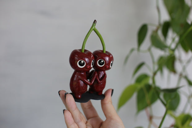 Sculpture of a pair of cherry characters, with large cute eyes and small mouths. They press their palms against one another and their heads are attached to the same stem.