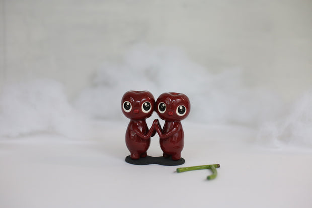 Sculpture of a pair of cherry characters, with large cute eyes and small mouths. They press their palms against one another and their stem sits nearby, removable. 