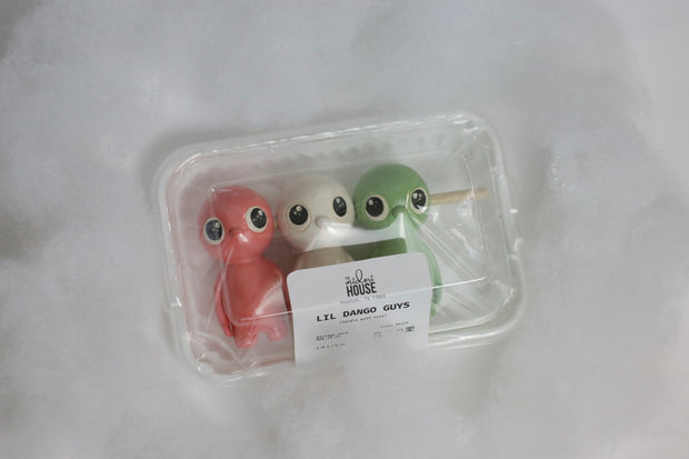 Sculpture of small characters shaped and colored to look like dango, with a stick going through their heads. They have cute eyes and a simple nose as their only facial feature. They are in product packaging, like they are being sold in a convenience store.