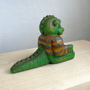 Painted whittled wooden sculpture of a baby Godzilla, with cute large eyes and a smiling sharp toothed expression. It sits on the ground with its arms out in front and wears a brown and blue striped sweater.