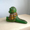 Painted whittled wooden sculpture of a baby Godzilla, with cute large eyes and a smiling sharp toothed expression. It sits on the ground with its arms out in front and wears a brown and blue striped sweater.