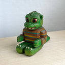 Painted whittled wooden sculpture of a baby Godzilla, with cute large eyes and a smiling sharp toothed expression. It sits on the ground with its arms out in front and wears a brown and blue striped sweater.