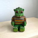 Painted whittled wooden sculpture of a baby Godzilla, with cute large eyes and a smiling sharp toothed expression. It sits on the ground with its arms out in front and wears a brown and blue striped sweater.