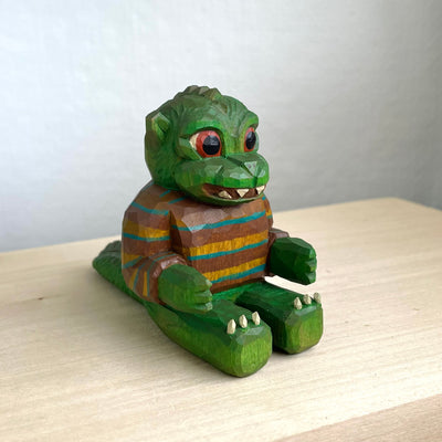 Painted whittled wooden sculpture of a baby Godzilla, with cute large eyes and a smiling sharp toothed expression. It sits on the ground with its arms out in front and wears a brown and blue striped sweater.