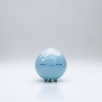 Small glossy blue ceramic sculpture of a round character with tiny legs and a closed eye peaceful expression. The top of its head is white like a snow capped mountain.