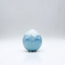 Small glossy blue ceramic sculpture of a round character with tiny legs and a closed eye slightly angry expression. The top of its head is white like a snow capped mountain.
