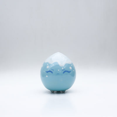 Small glossy blue ceramic sculpture of a round character with tiny legs and a closed eye slightly angry expression. The top of its head is white like a snow capped mountain.