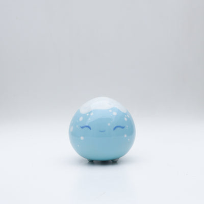 Small glossy blue ceramic sculpture of a round character with tiny legs and a closed eye gleeful expression. The top of its head is white like a snow capped mountain.