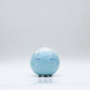 Small glossy blue ceramic sculpture of a round character with tiny legs and a closed eye gleeful expression. The top of its head is white like a snow capped mountain.