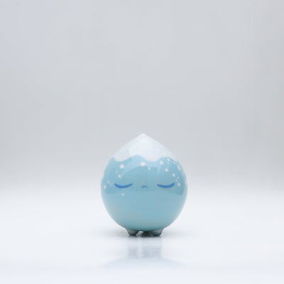Small glossy blue ceramic sculpture of a round character with tiny legs and a closed eye, slightly pouty expression. The top of its head is white like a snow capped mountain.