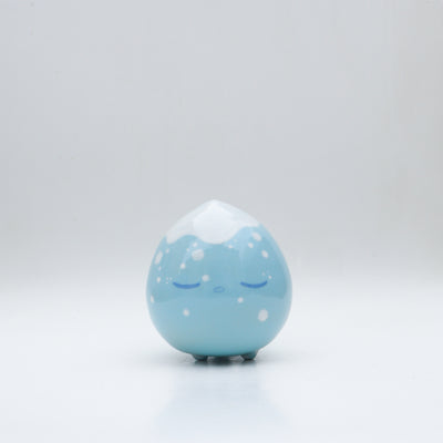 Small glossy blue ceramic sculpture of a round character with tiny legs and a closed eye, sleepy expression. The top of its head is white like a snow capped mountain.
