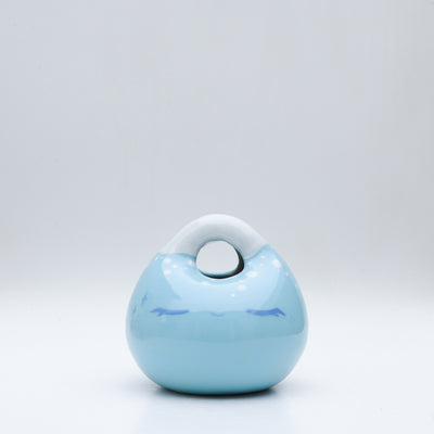 Ceramic sculpture made to look like a smooth handbag, blue with a cute cartoon face on it. It has a closed eye expression with a small smile and white dots along the top, like a snowcapped mountain.