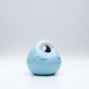 Ceramic sculpture made to look like a smooth handbag, blue with a cute cartoon face on it. It has a closed eye expression with a small smile and white dots along the top, like a snowcapped mountain.