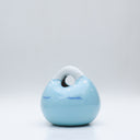 Ceramic sculpture made to look like a smooth handbag, blue with a cute cartoon face on it. It has a closed eye expression with a small smile and white dots along the top, like a snowcapped mountain.