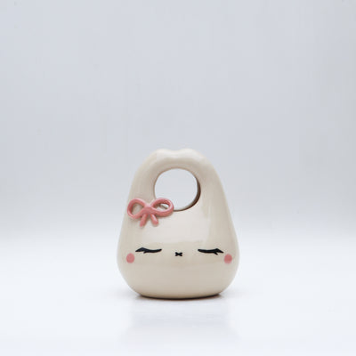 Ceramic sculpture made to look like a purse, with a cute closed eye bunny face and rosy cheeks. It has a pink bow atop its head.