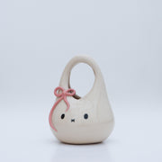 Ceramic sculpture made to look like a purse, with the face of Miffy the bunny, dot eyes and an 'x' for a mouth. It has a pink bow atop its head.
