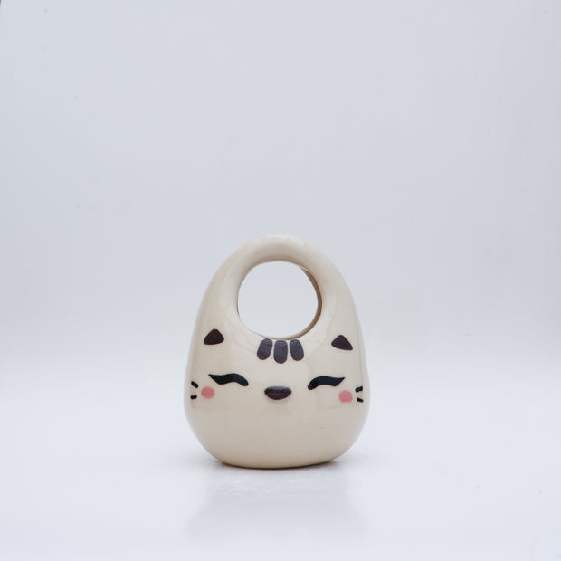 Ceramic sculpture made to look like a purse, with the face of a cute, closed eye cat with rosy cheeks and 3 stripes at the top of its head.
