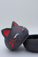 Black clay pot with a lid that looks like a fox's head, a matching black color with red ears and face paint. It has closed eyes and a small white nose. The bowl has no body details.