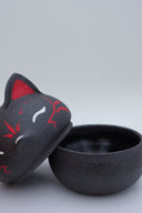 Black clay pot with a lid that looks like a fox's head, a matching black color with red ears and face paint. It has closed eyes and a small white nose. The bowl has no body details.