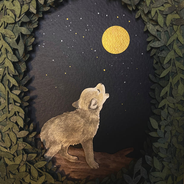 Framed paper cutting artwork of a small wolf, standing and howling up at the moon, gold in the dark night sky. Piece is framed by an oval assembling of cut leaves.