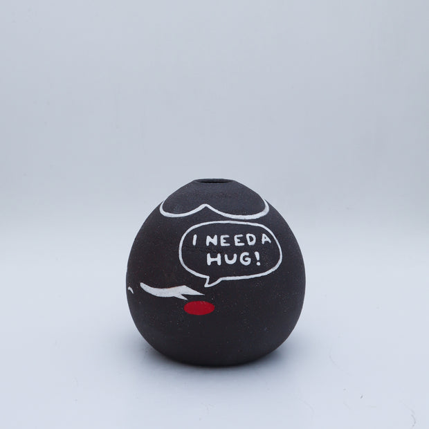 Small black vase with a drawing of a character with closed eyes and rosy cheeks. They have a speech bubble that reads "I Need a Hug!"