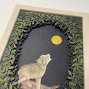 Framed paper cutting artwork of a small wolf, standing and howling up at the moon, gold in the dark night sky. Piece is framed by an oval assembling of cut leaves.