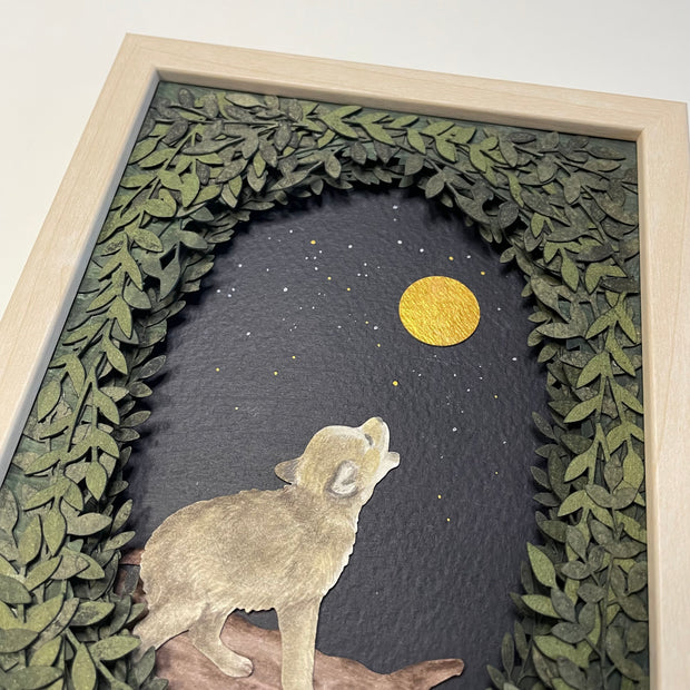 Framed paper cutting artwork of a small wolf, standing and howling up at the moon, gold in the dark night sky. Piece is framed by an oval assembling of cut leaves.