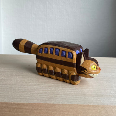 Whittled wooden sculpture of a cat shaped like a bus from My Neighbor Totoro. Its tail sticks straight out and it has a smiling face.