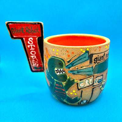 Ceramic mug with brightly painted graphics and a slab that juts out, that reads "Giant Robot Store." Drawings show a cute green Godzilla roaring at GR2 Gallery.