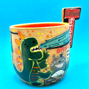 Ceramic mug with brightly painted graphics and a slab that juts out, that reads "Giant Robot Store." Drawings show a large green Godzilla roaring at a store display of Totoro.