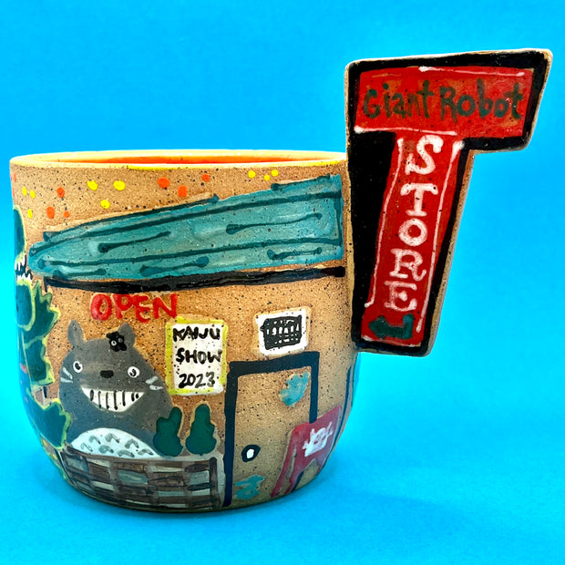 Ceramic mug with brightly painted graphics and a slab that juts out, that reads "Giant Robot Store." Drawings show Giant Robot Store's front window display, with a large Totoro cut out and a sign for the kaiju show 2023.