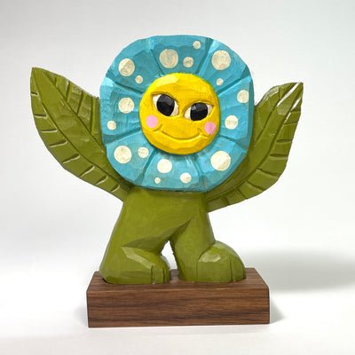 Painted whittled wooden sculpture of a smiling blue flower, with a yellow face and pink cheeks. Its body is green, with leaves for arms and stumpy feet.