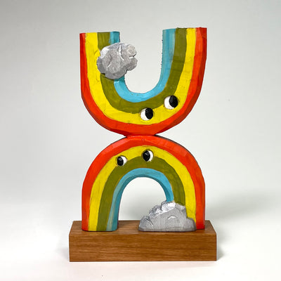 Painted whittled wood sculpture of 2 rainbows, one upside down and stuck to the top of the other. The have eyes and a single silver cloud attached to each.