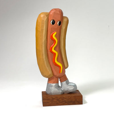 Painted whittled wood sculpture of a hot dog, standing upright and wearing a pair of silver shoes. It has eyes and no other features and squiggly mustard and ketchup lines.  