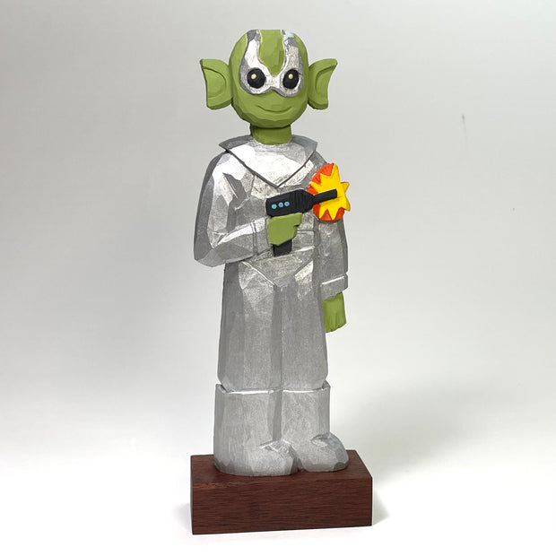 Painted whittled wood sculpture of a green alien, wearing a silver space suit and holding a laser gun, that emits a yellow and orange blast. The alien smiles.