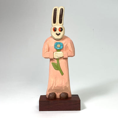 Painted whittled wood sculpture of a white bunny with red eyes and a heart shaped nose. It stands and wears a pink nightgown while holding a blue flower in its hands.