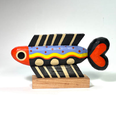Painted whittled wood sculpture of a fish, with a long skinny body and a heart shaped tail fin. Its fins are tan and black striped and its body is red, yellow, blue and black.